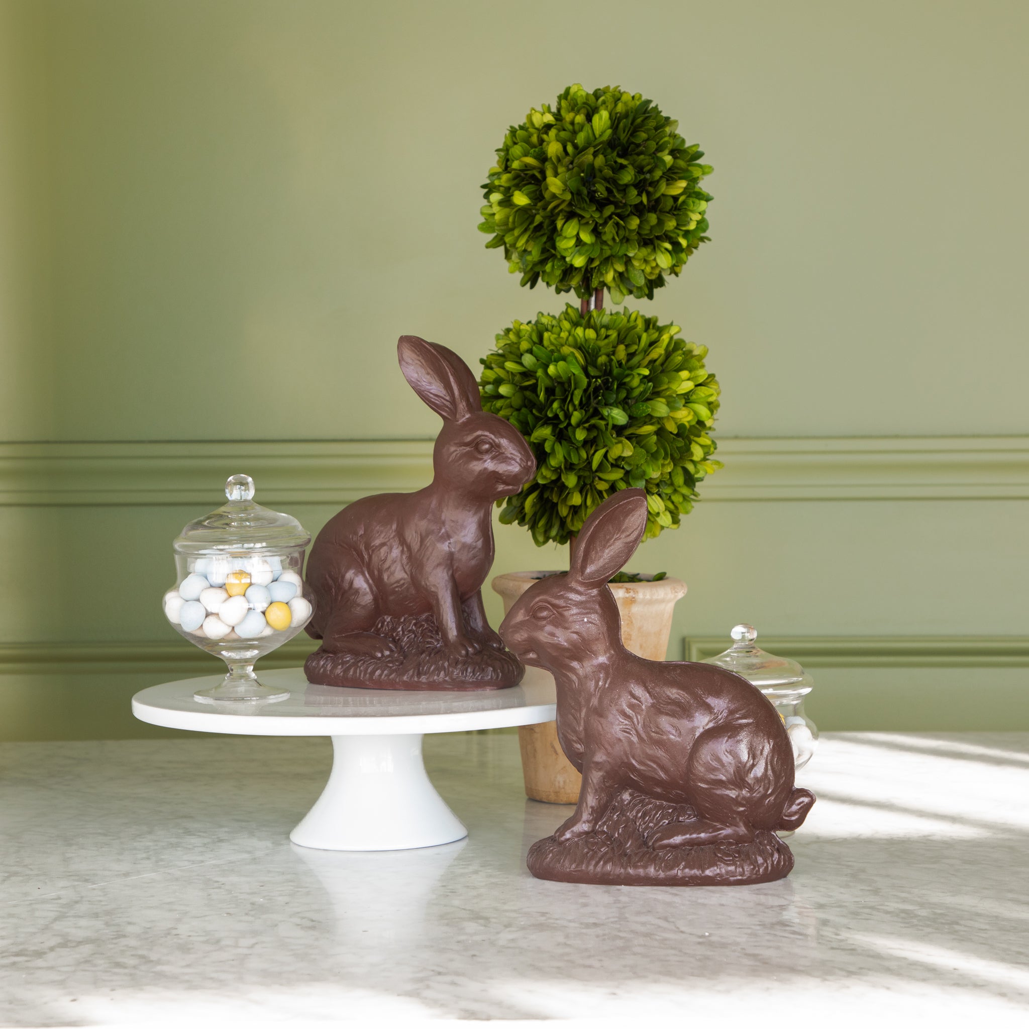 &quot;Chocolate&quot; Sitting Bunnies displayed with candy eggs in a jar and topiary in the background.