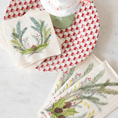 A top-down view of a table with a stack of Christmas Sprigs Cocktail Napkins and a stack of Christmas Sprigs Guest Napkins, fanned out next to a red and white plate and a cup of hot chocolate.