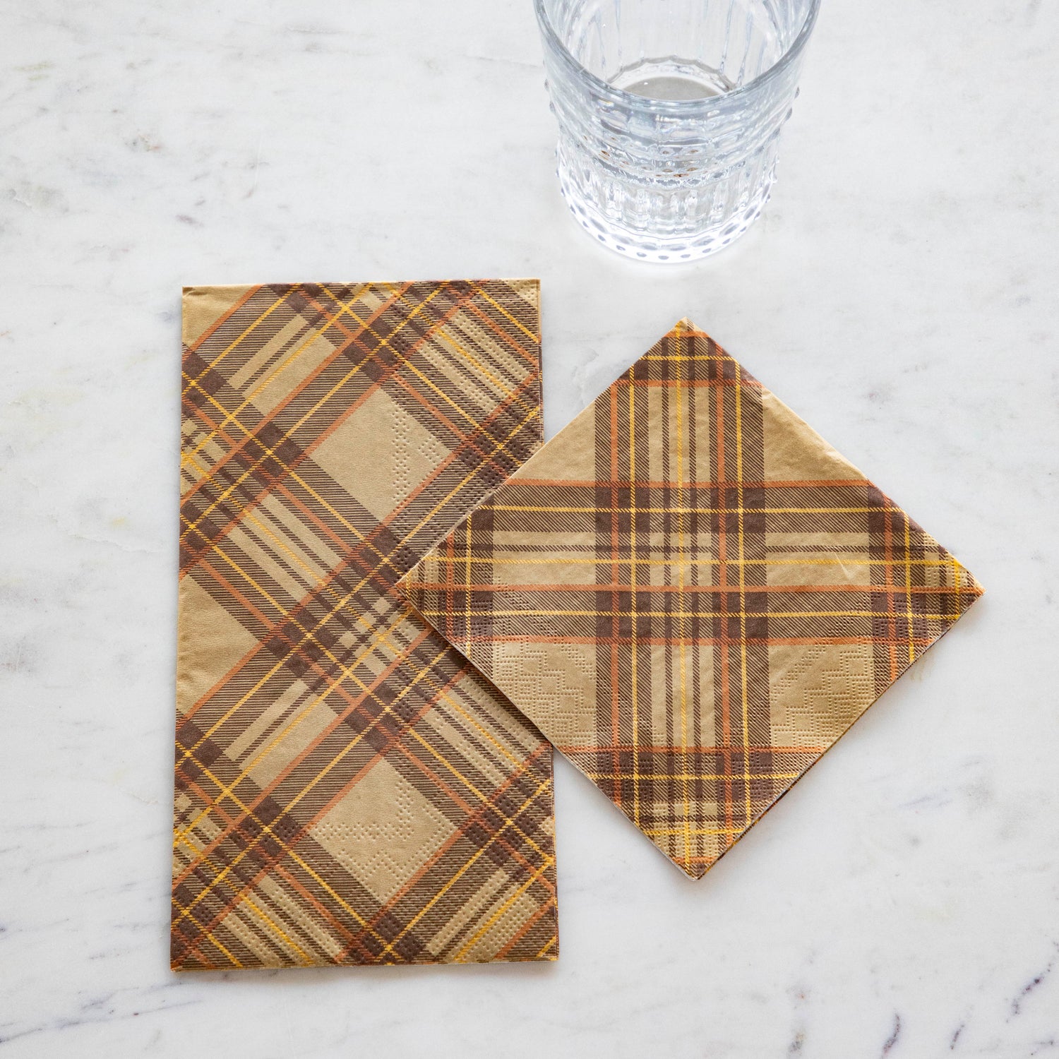 An Autumn Plaid Guest Napkin and Cocktail Napkin on a white marble table next to a clear glass.