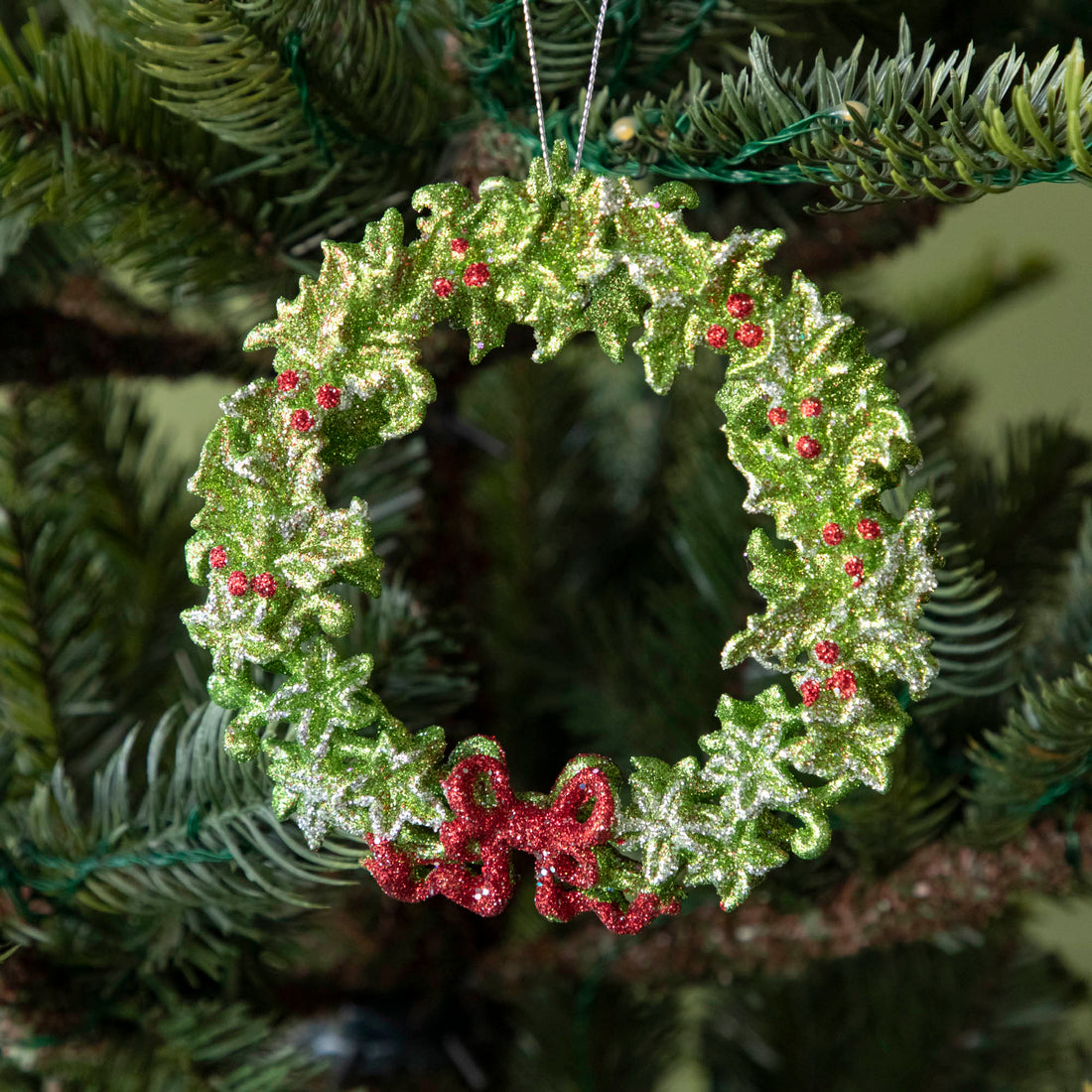 A 5&quot; acrylic green wreath ornament with silver accents, bright red berries and a red bow, hanging on a tree by a silver string.