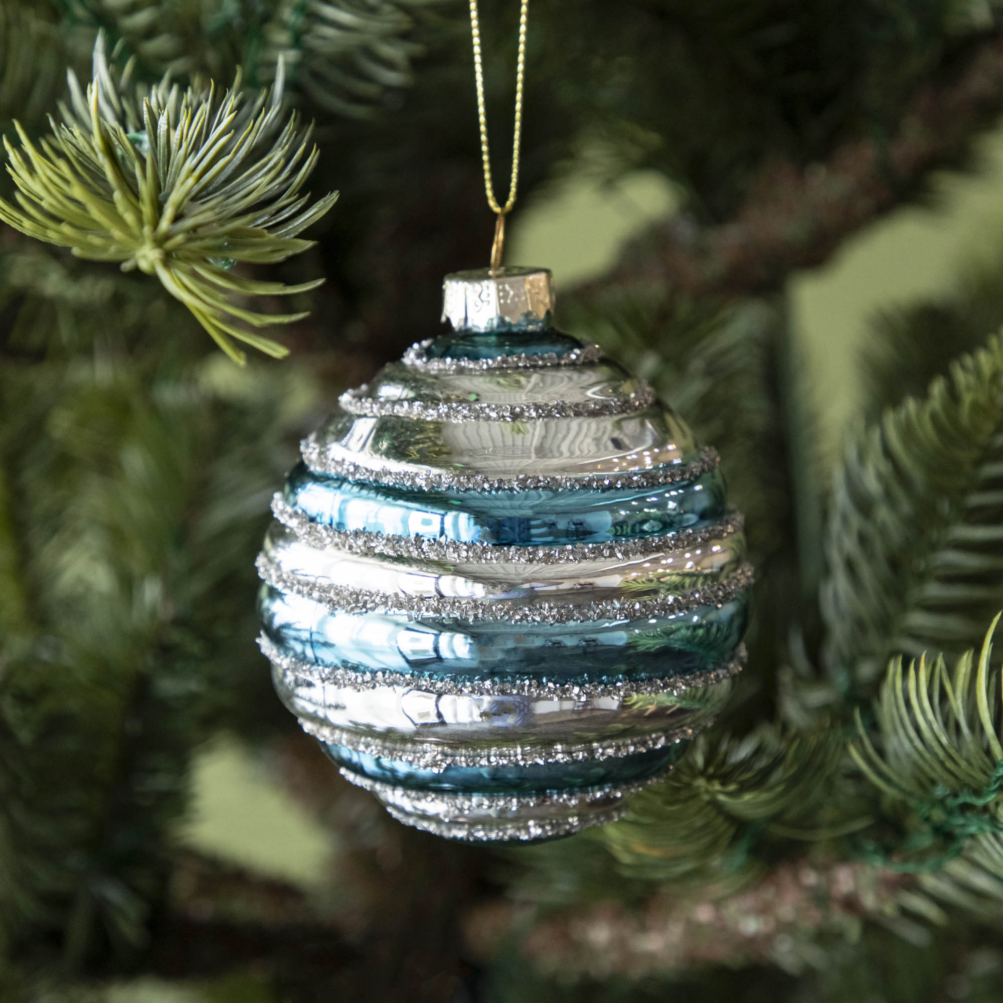 A Glass Silver Stripe Ball Ornament with blue stripes and silver glitter, hanging on a tree by a gold string.