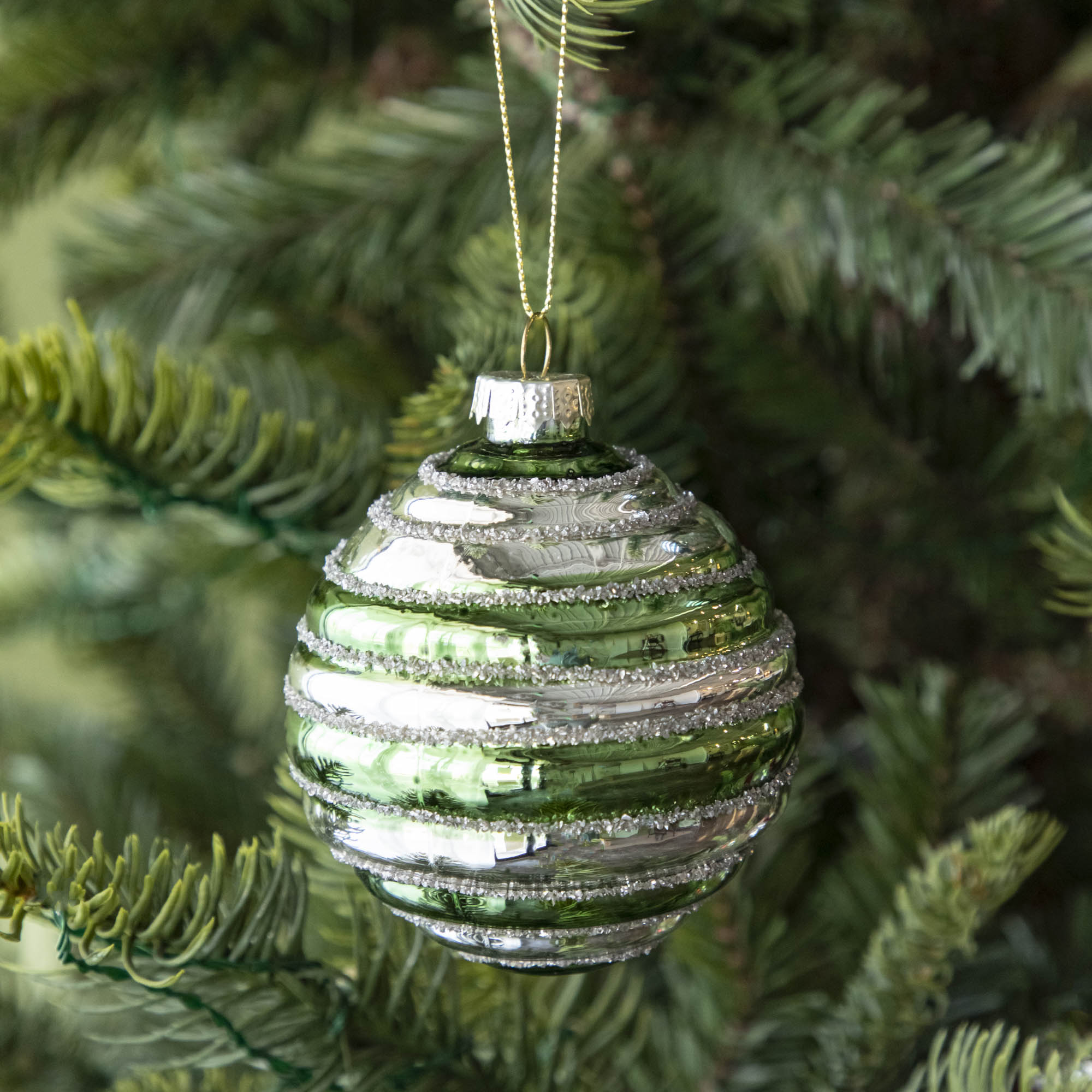 A Glass Silver Stripe Ball Ornament with green stripes and silver glitter, hanging on a tree by a gold string.