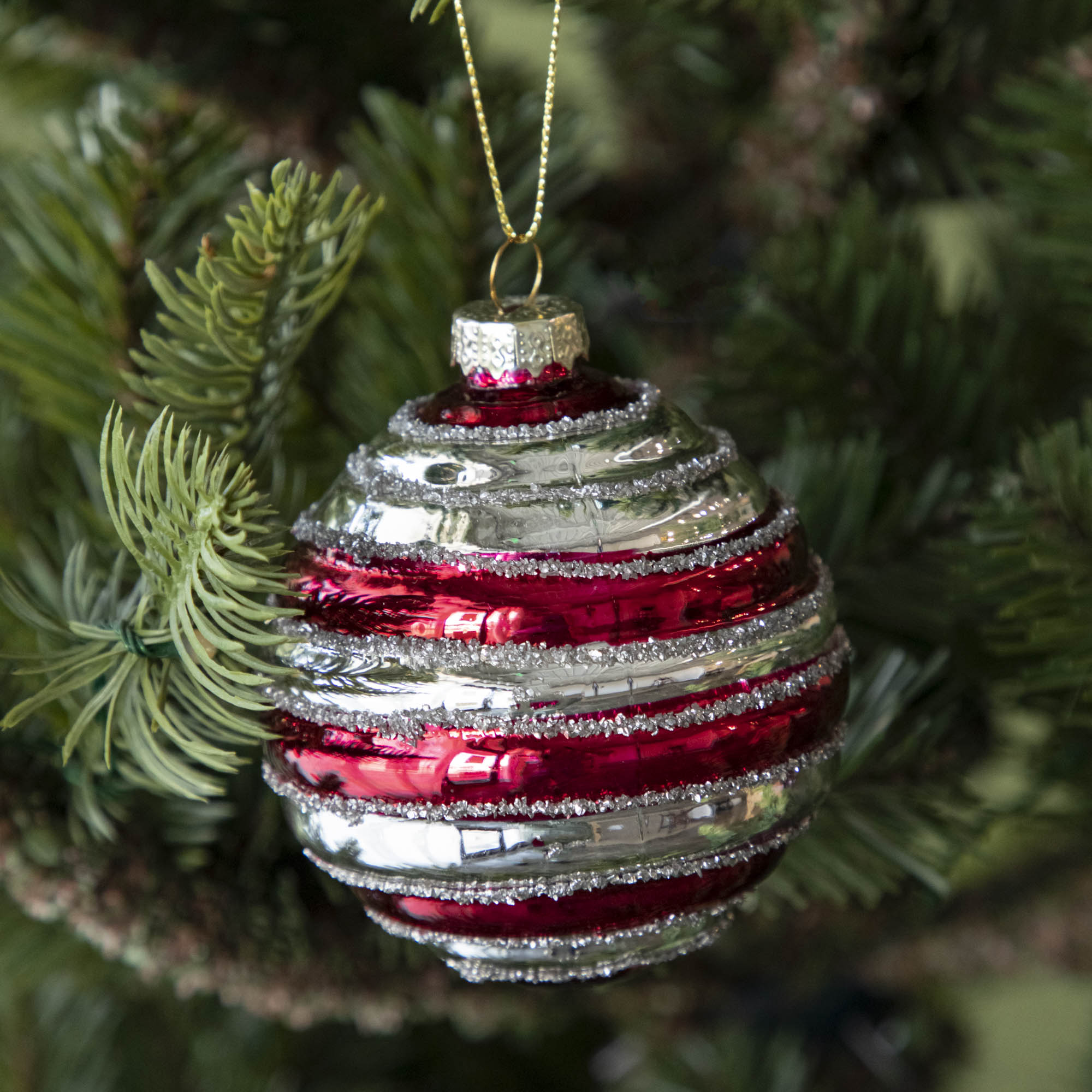 A Glass Silver Stripe Ball Ornament with red stripes and silver glitter, hanging on a tree by a gold string.