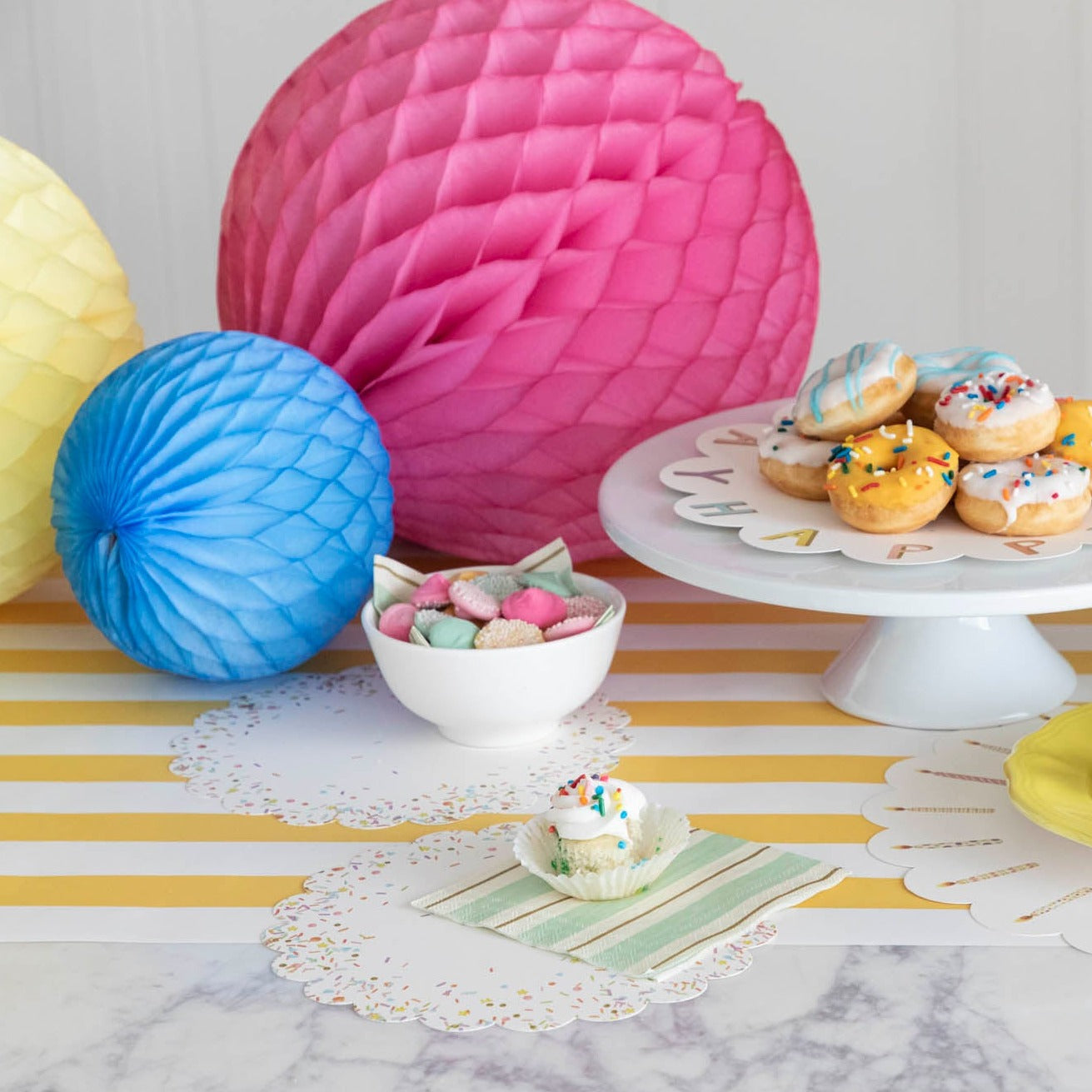 A table adorned with Happy Birthday Serving Papers from Hester &amp; Cook and charcuterie spreads, featuring an assortment of paper balls and donuts.