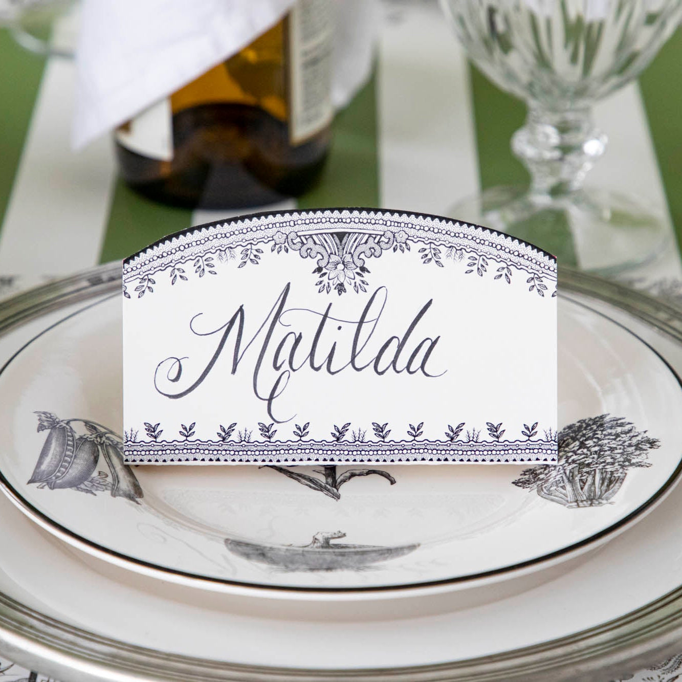 Black Willow Place Card with &quot;Matilda&quot; written on it, resting on dinner plates on a place setting.