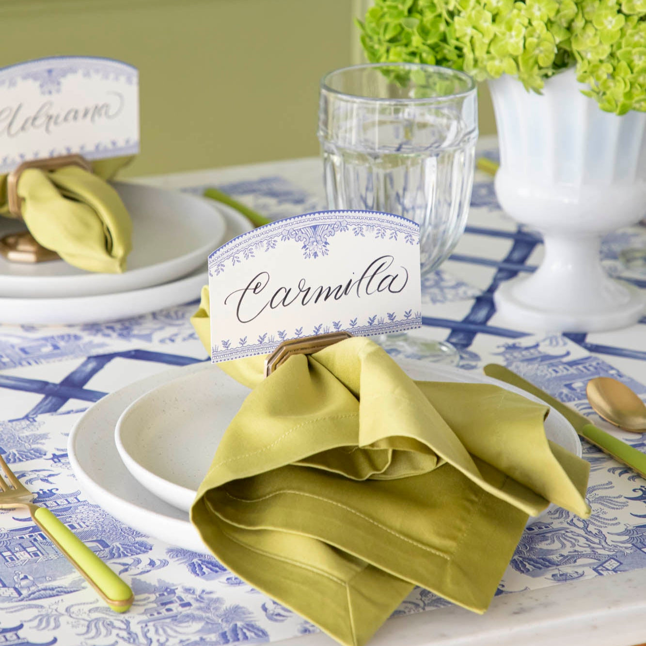 The Blue Willow Place card with &quot;Carmilla&quot; written in the white space, held up by the Brass Napkin Ring with Place Card Holder, on an elegant table setting for two.