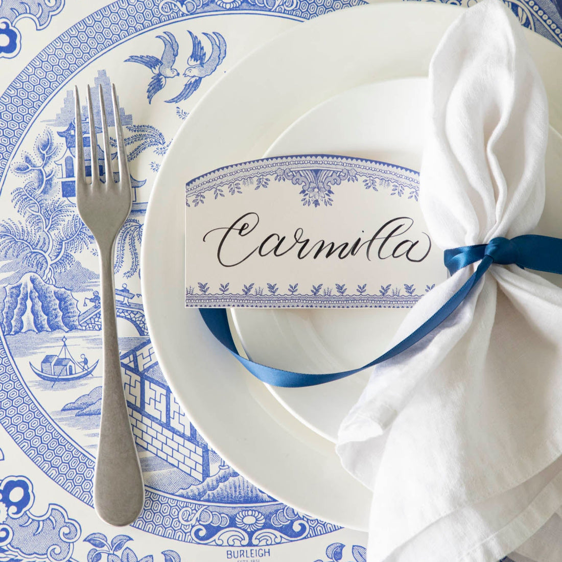 Burleigh Blue Place Card with &quot;Camilla&quot; written in the white space, resting on dinner plates, on top of the Die-cut Blue Willow Plate Placemat. 