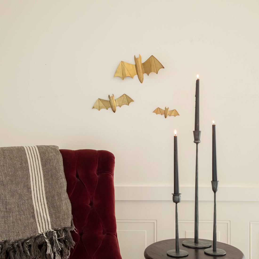 Metal winged bats on a wall with a burgundy chair and black taper candles on a table.