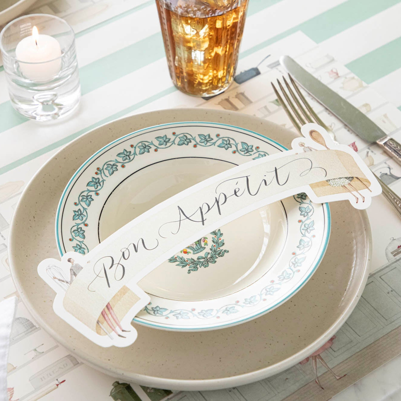 Two Cooks Banner Table Accent with &quot;Bon Appetit&quot; written on it, resting on dinner plates, on an elegant place setting for one.