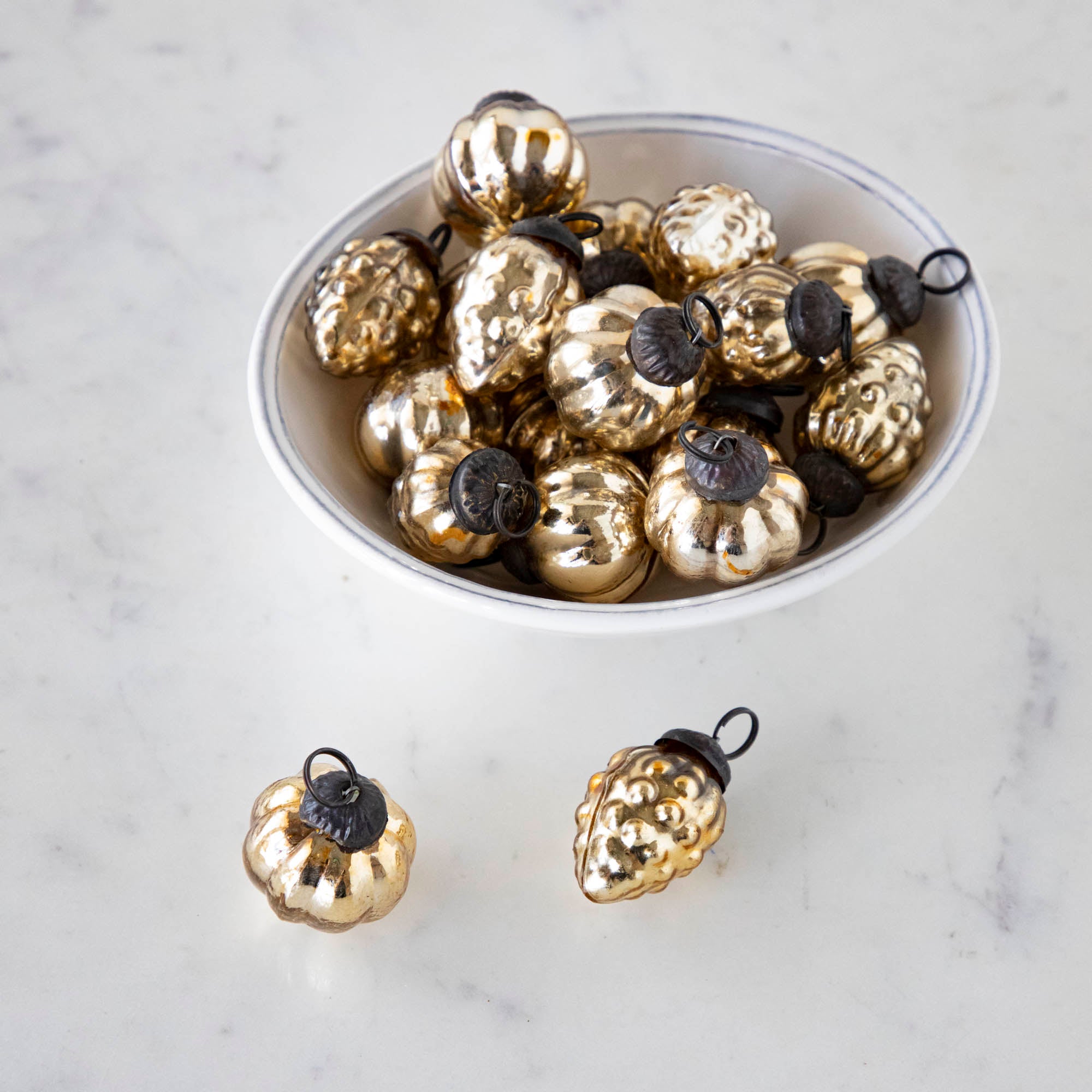 A white bowl on a marble table filled with one inch Embossed Antique Gold Mercury Glass Ornaments and two outside of the bowl.