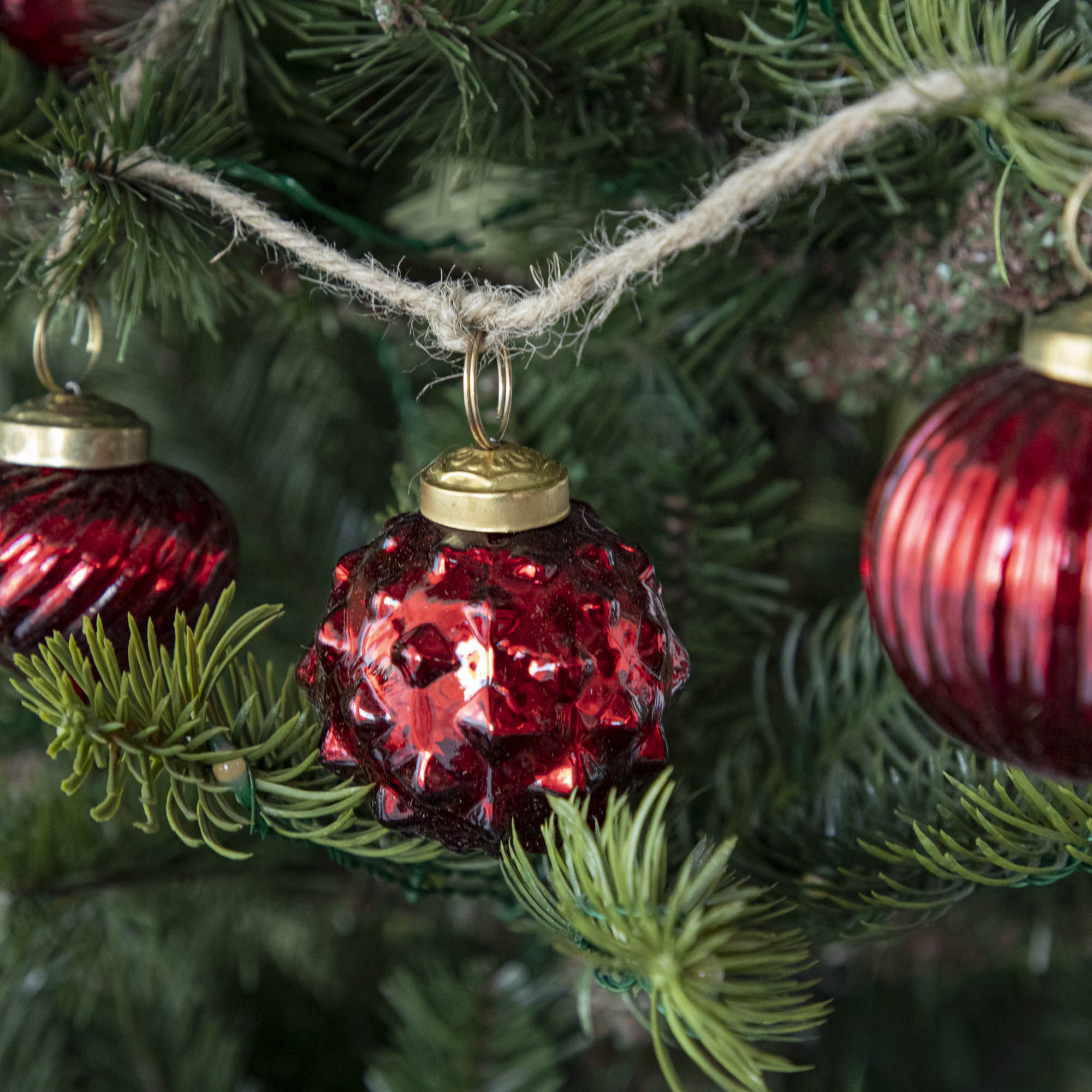 A close up of three Embossed Red Mercury Glass Ornaments showing the detailing and jute cord hanging on a tree.