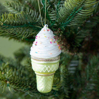 An Ice Cream Cone ornament with a sparkled green cone, white icing and sprinkles, hanging on a tree by a green string.