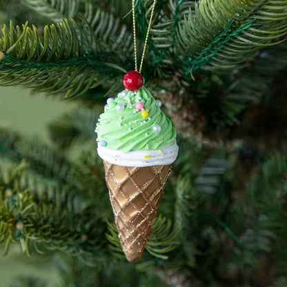 An Ice Cream Cone ornament with a sparkled brown cone, green and white icing and sprinkles with a cherry on top, hanging on a tree by a green string.