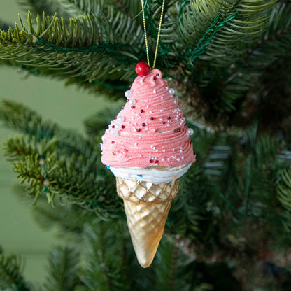 An Ice Cream Cone ornament with a sparkled tan cone, pink and white white icing with sprinkles and a cherry on top, hanging on a tree by a green string.