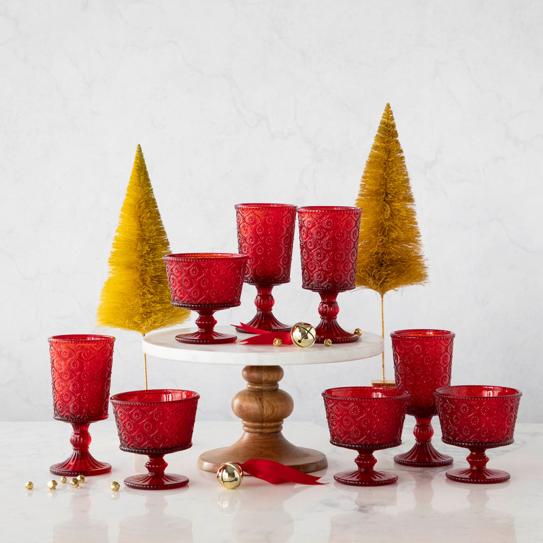 Elegant Qualia red pressed glassware set displayed on a white cake stand with wooden pedestal, surrounded by gold flocked trees and scattered jingle bell ornaments.