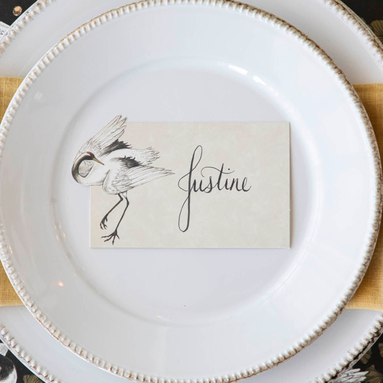 Crane Dance Place Card with &quot;Justine&quot; written on it, laying flat on a set of dinner plates.