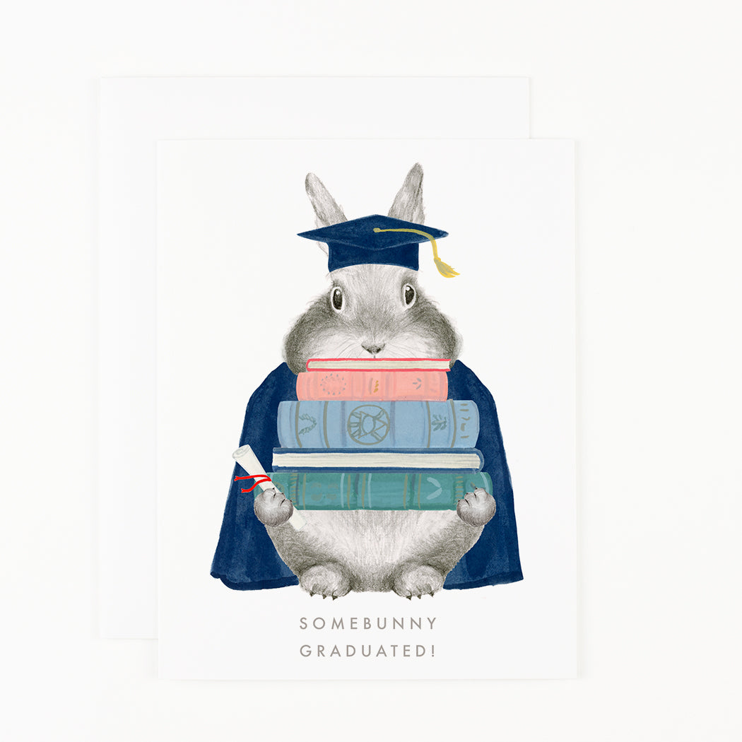 A Somebunny Graduated Card from Dear Hancock, featuring a charming bunny adorned in a cap and gown, clutching books tightly.