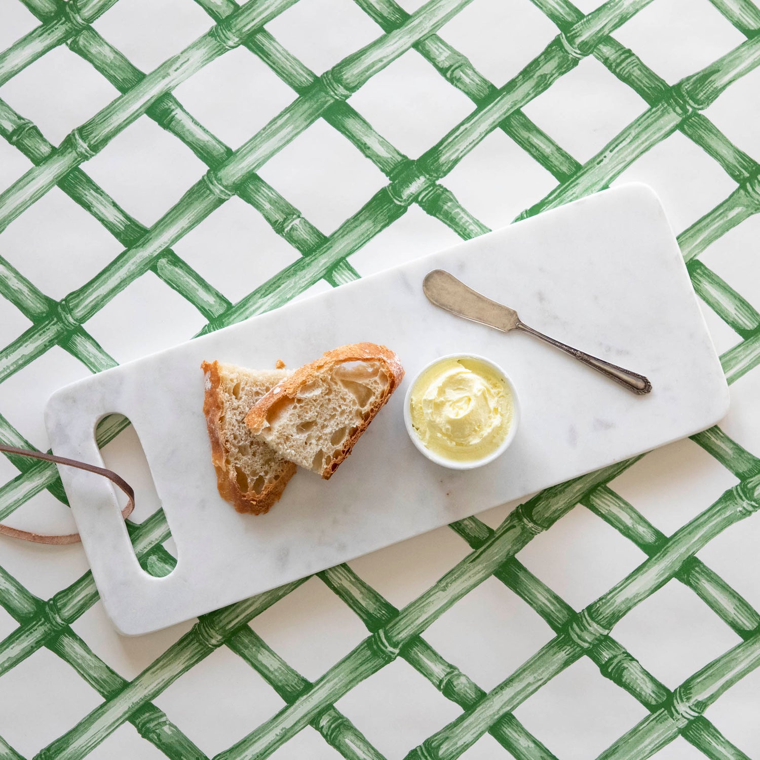 A White Marble Cheese Board with sliced bread and butter and a butter spreader on top, on top of a green lattice runner.