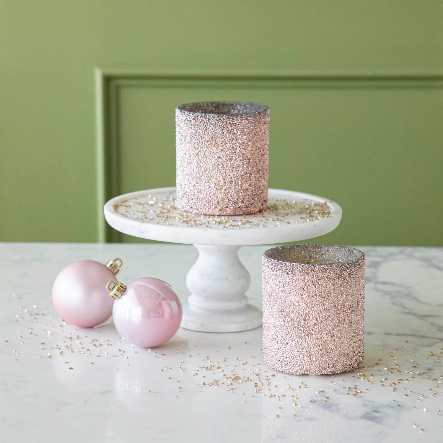 Pink Shimmer Votives styled with one on a cake stand and one on the table, surrounded by glitter and two pink ornaments.