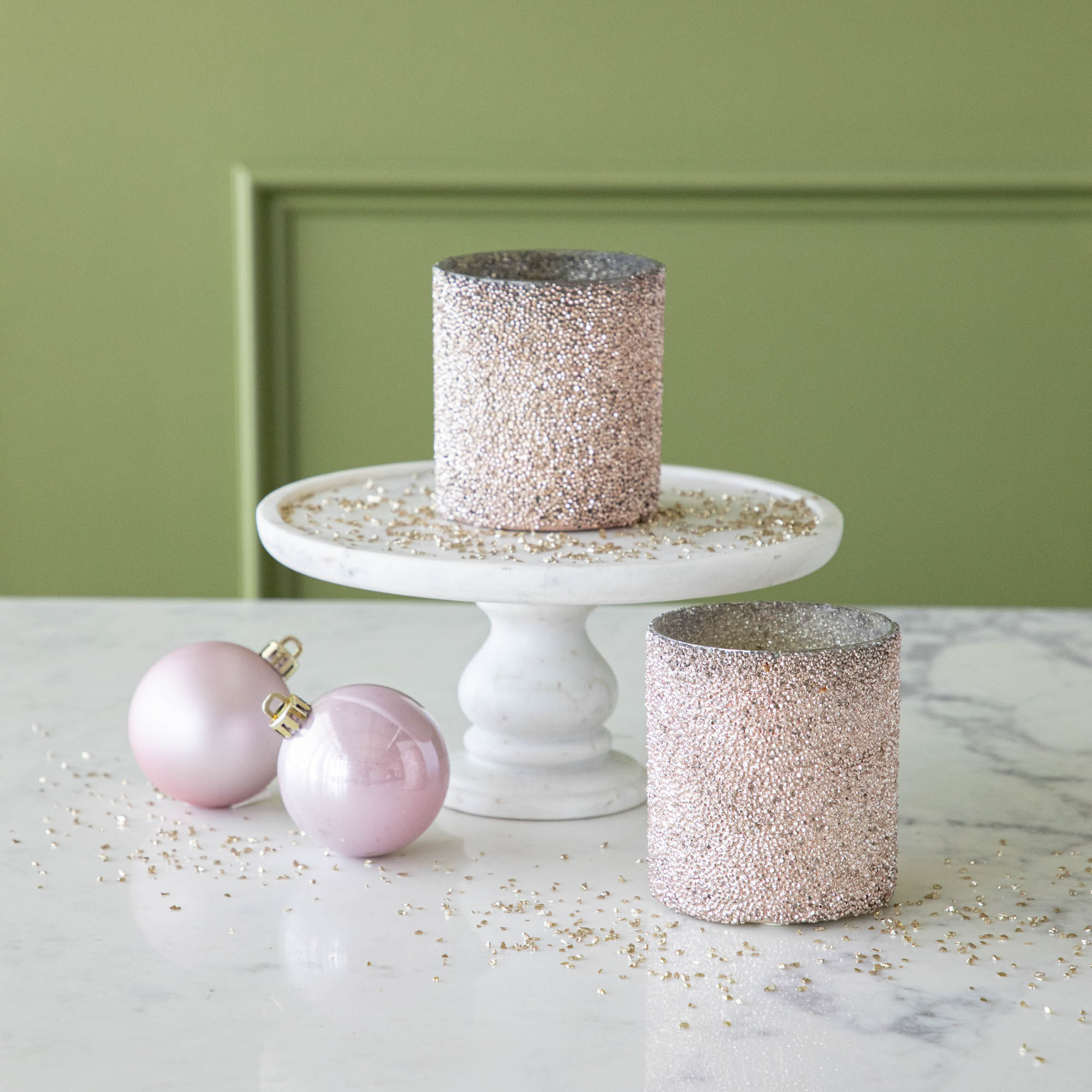 Pink Shimmer Votives styled with one on a cake stand and one on the table, surrounded by glitter and two pink ornaments.