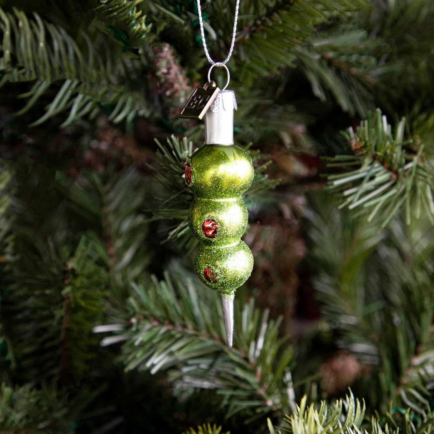 The Three Olives Please Ornament hanging on a tree.