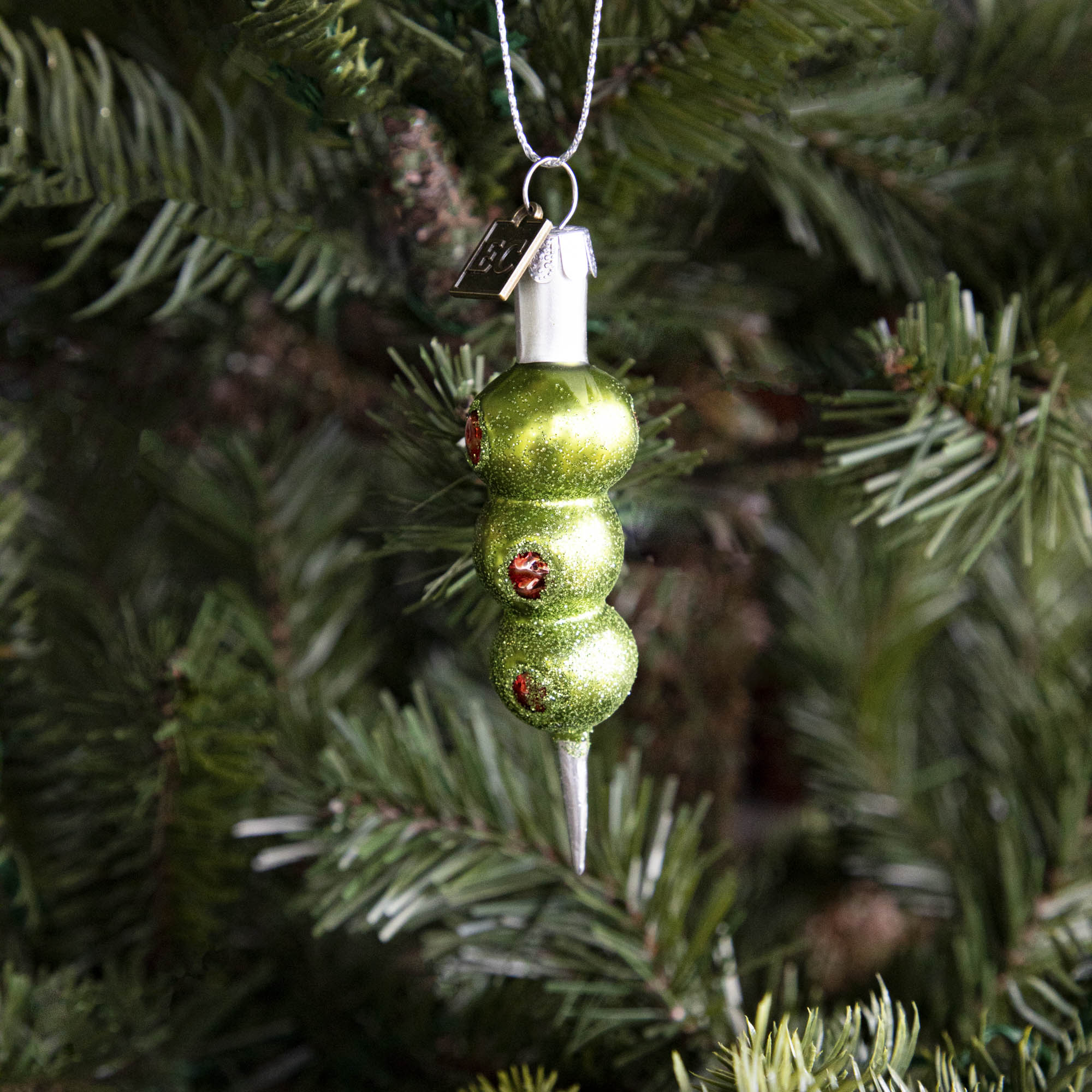 The Three Olives Please Ornament hanging on a tree.