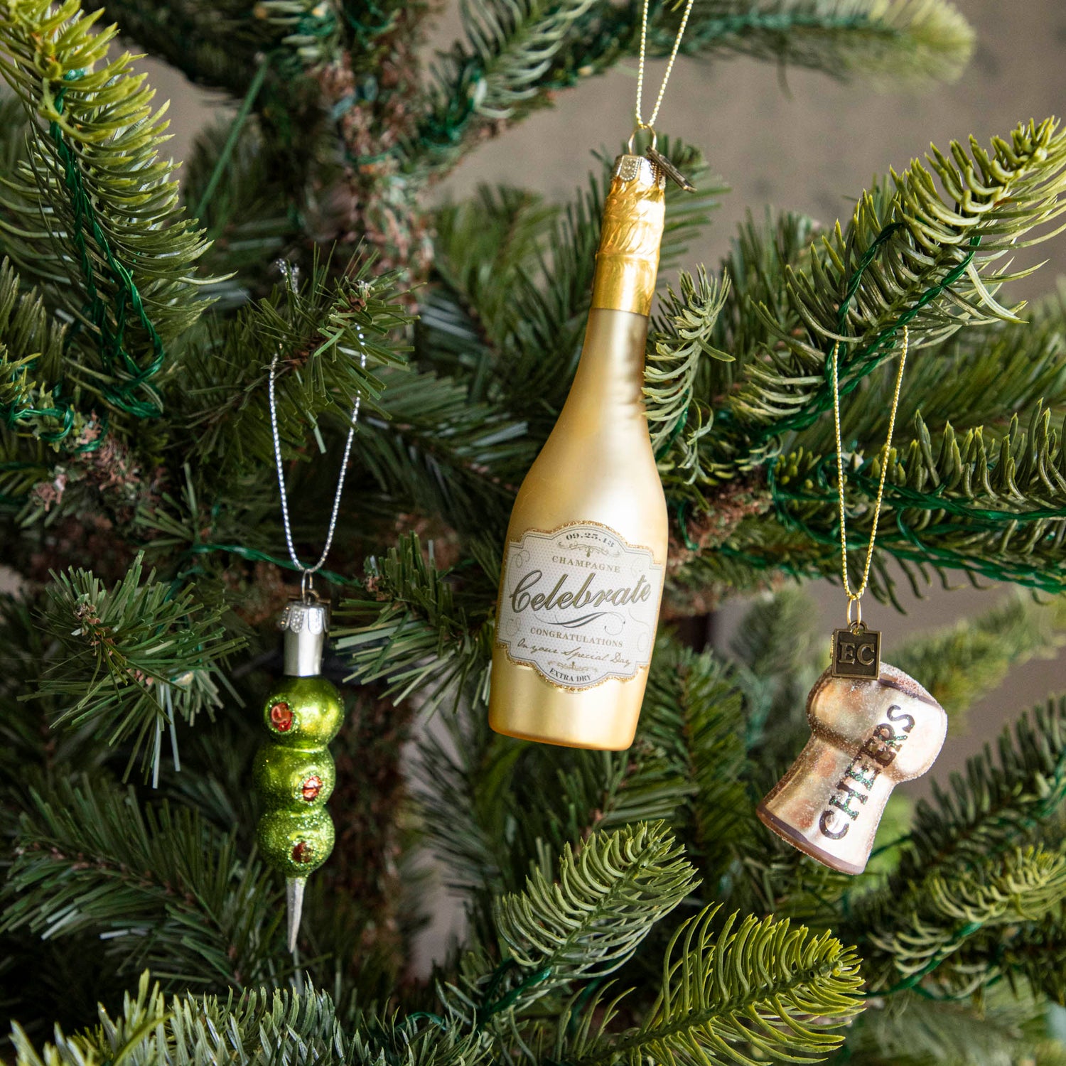 Congratulations Gold Champagne Ornament, Three Olives Please Ornament and Cheers Cork Ornament hanging on a tree.