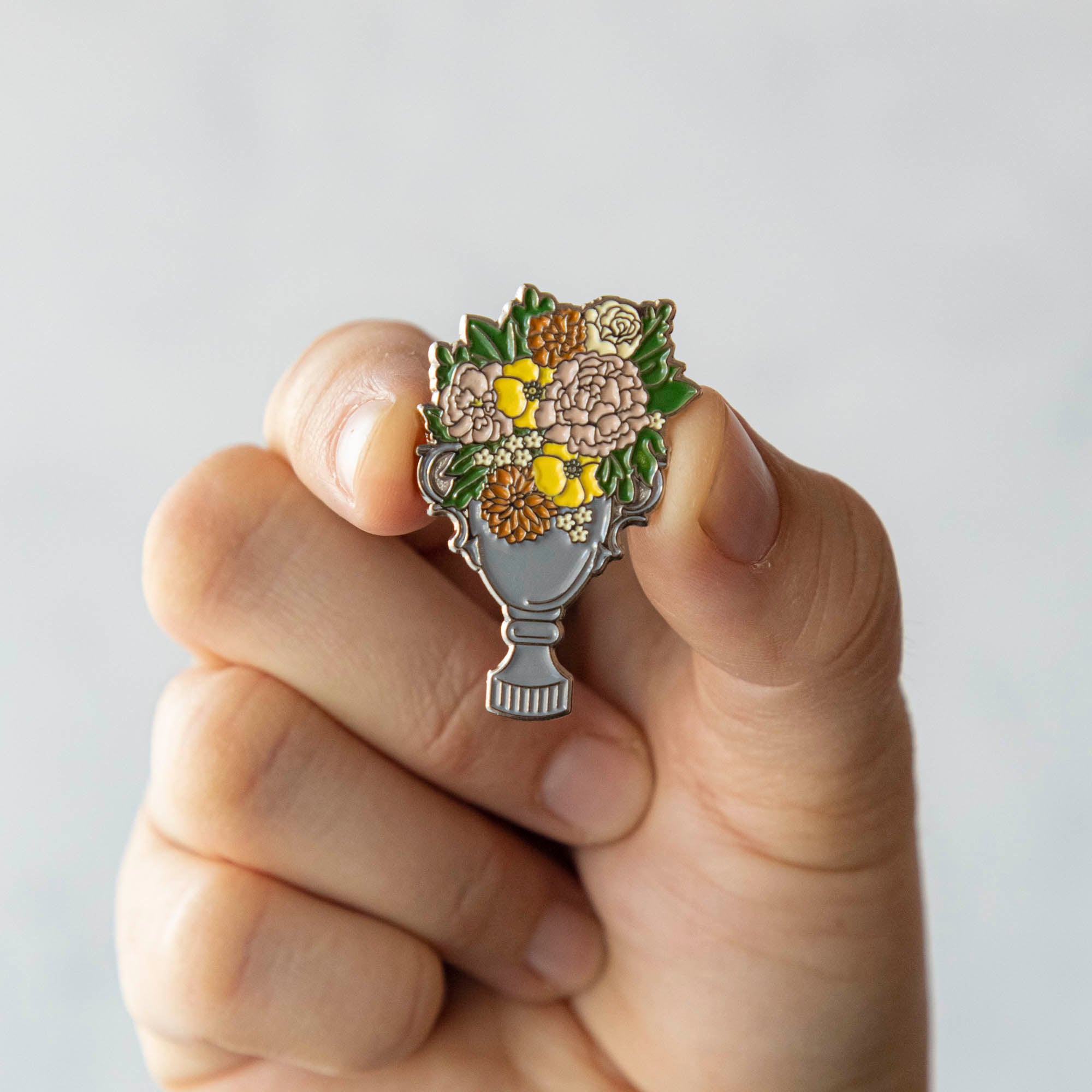 An Hester &amp; Cook enamel pin depicting a garden trophy with flowers in a vase, perfect for decorating a backpack.