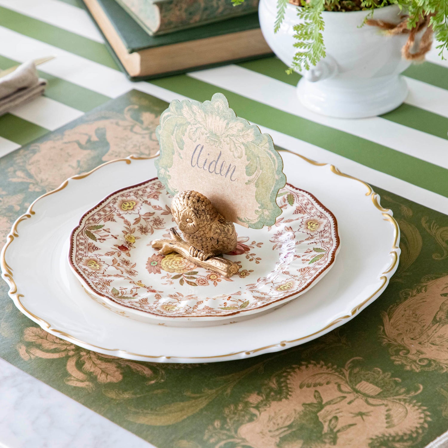 The Owl Name Card Holder holding the Moss Fable Toile Place Card with &quot;Aiden&quot; written on it in cursive, resting on two dinner plates with the Moss Fable Toile Placemat and Moss Green Classic Stripe Runner underneath.