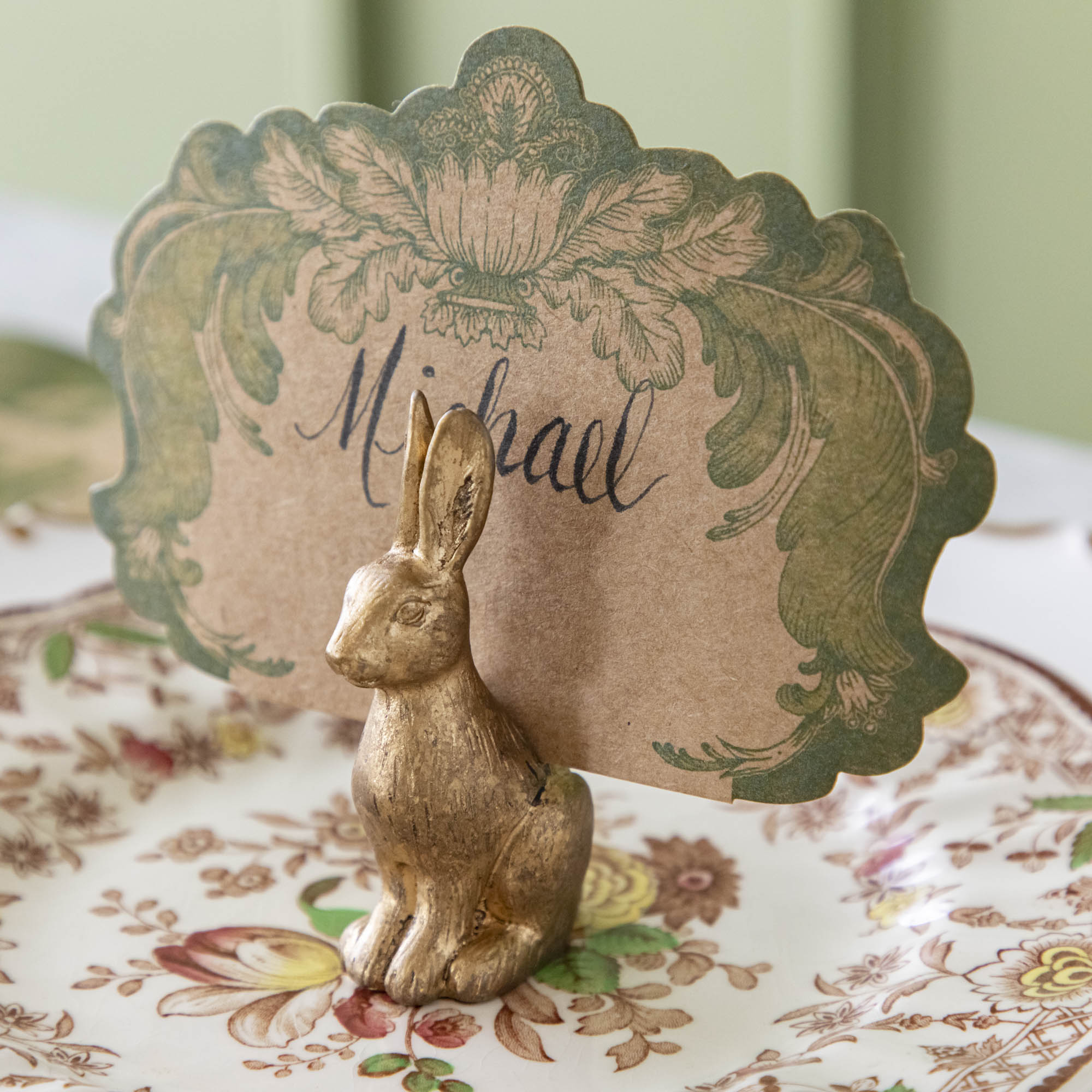 The antique gold finish Rabbit Name Card holder, holding the Moss Fable Toile Place Card with &quot;Michael&quot; written in cursive, sitting on a floral plate.