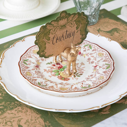 The Fox Name Card Holder holding the Moss Fable Toile Place Card with &quot;Courtney&quot; written in cursive, standing on two dinner plates with the Moss Fable Toile Placemat and Moss Green Classic Stripe Runner underneath.