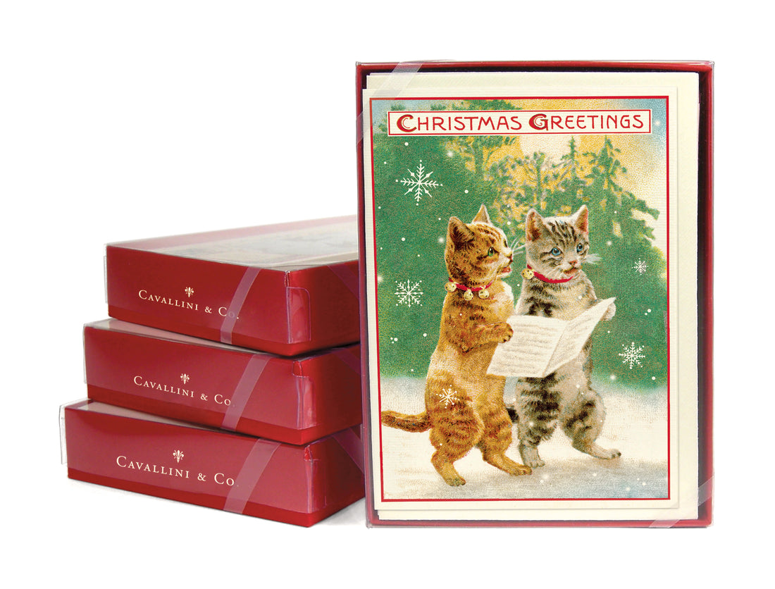 Three stacked card sets next to a front facing boxed card set of the Christmas Cats depicting two cats holding a paper in a snowy woods scene and  &quot;Christmas Greetings&quot; at the top of the card.