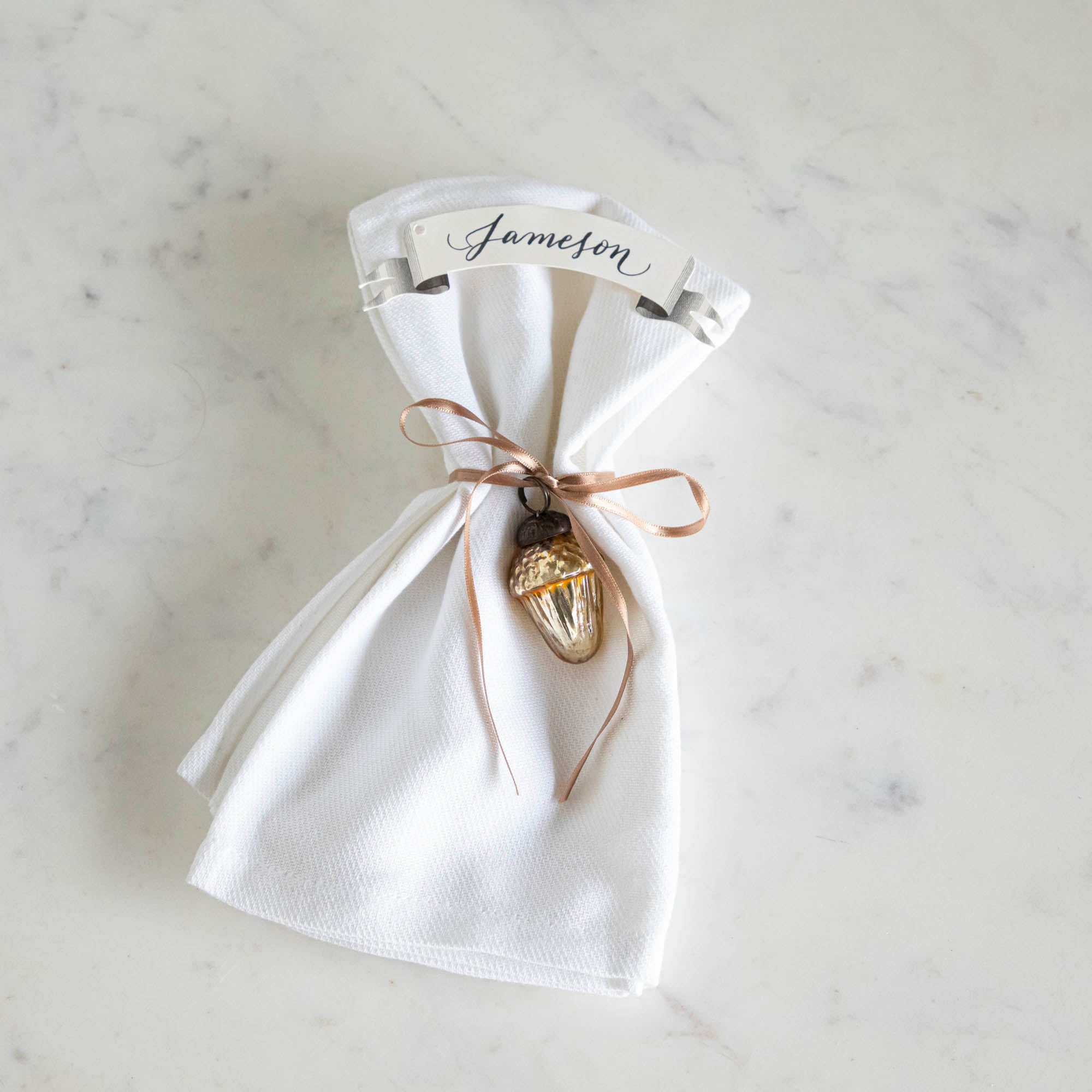 Glass acorn ornament tied around a napkin with a place card with the name &quot;Jameson&quot; written on it.