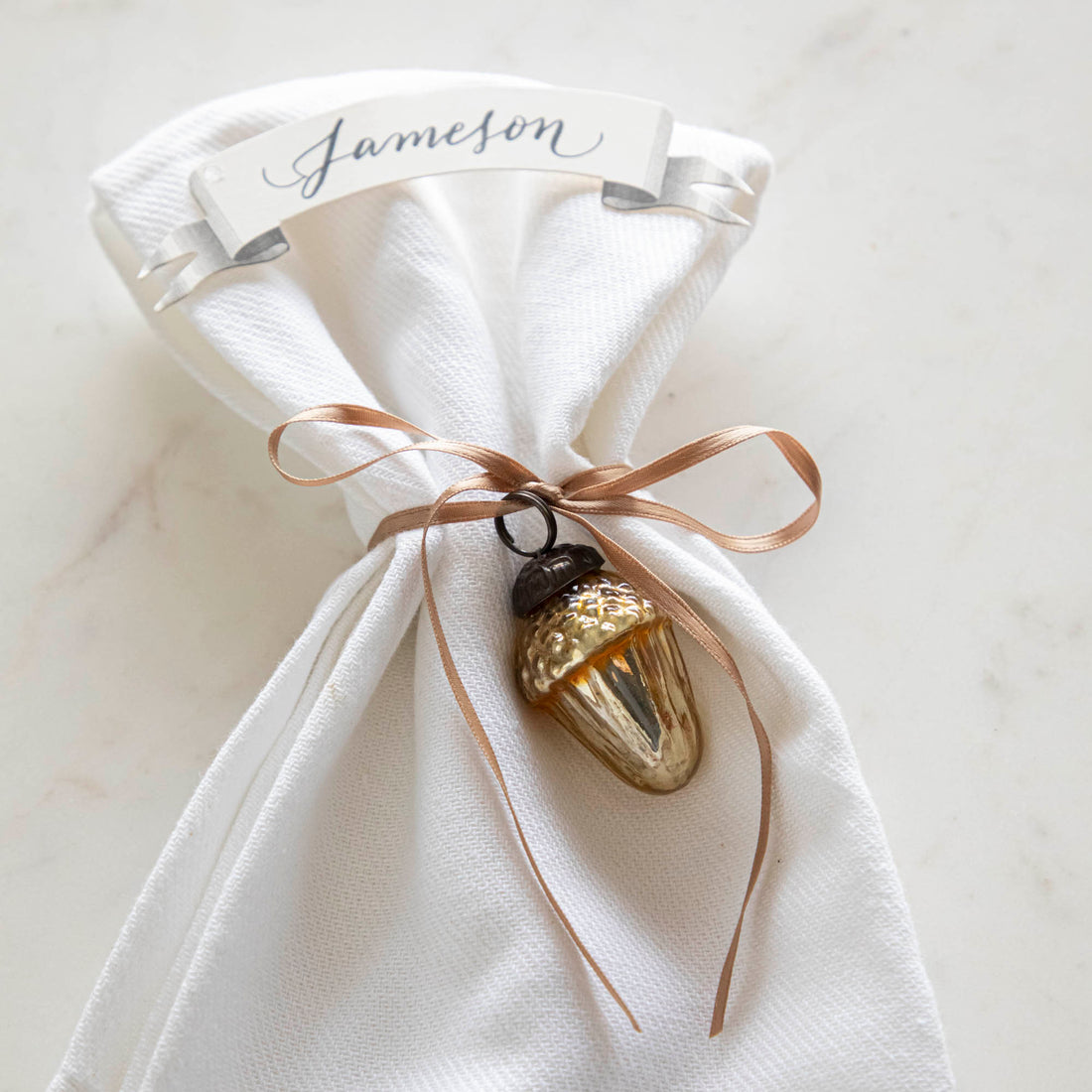 Glass acorn ornament tied around a napkin with a place card with the name &quot;Jameson&quot; written on it.