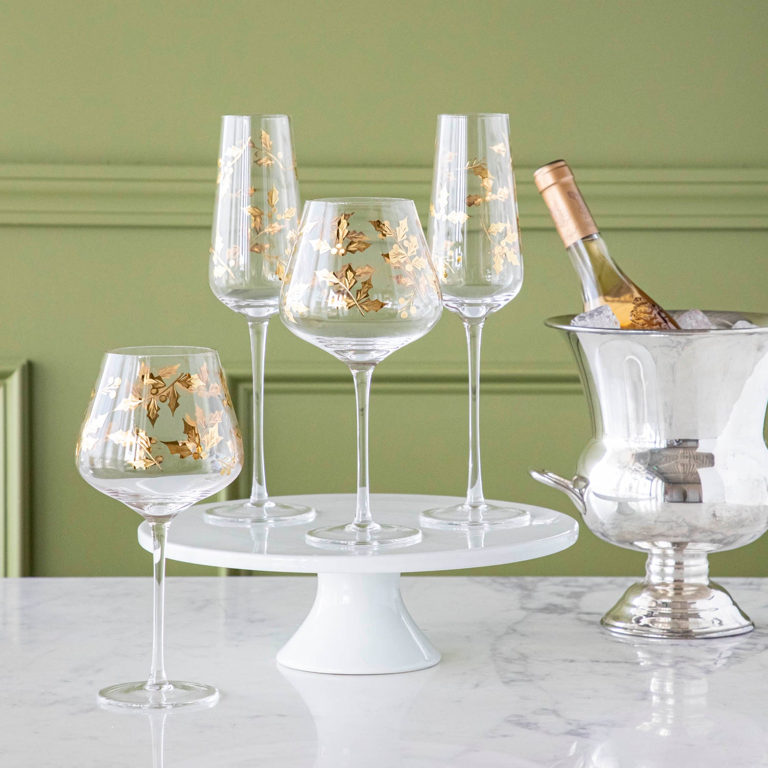 Gold Holly Etched Glassware on a cake stand and marble table, with a bottle of champagne in an ice bucket.