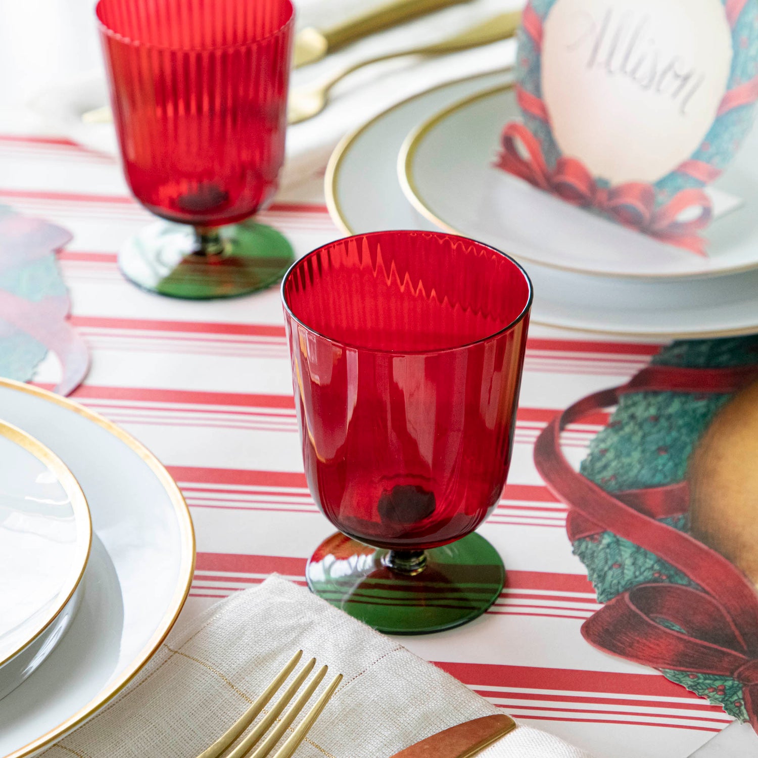 A green &amp; red stemmed flute glass on a red &amp; white stripe runner with gold rimmed plates.