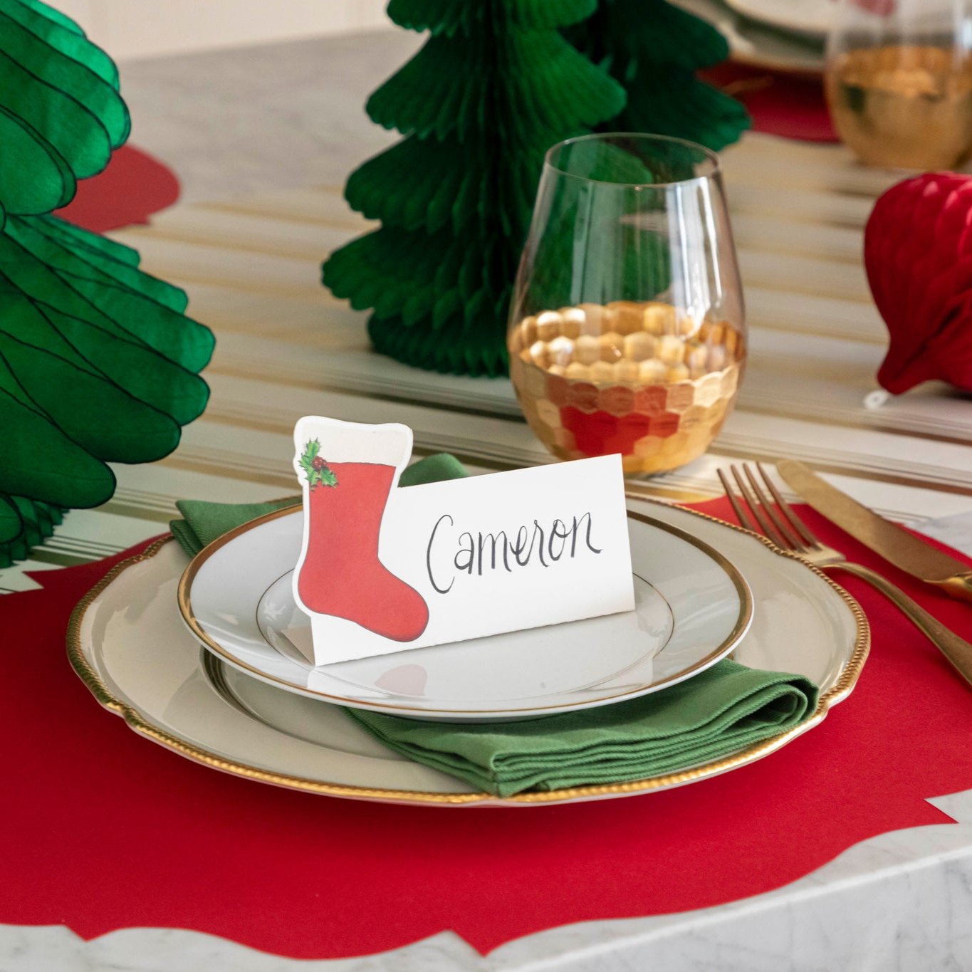 An elegant Christmas themed table setting with the Die-cut Red French Frame Placemat under gold rimmed plates, a green napkin, and a Stocking Place Card with the name &quot;Cameron&quot; written on it.