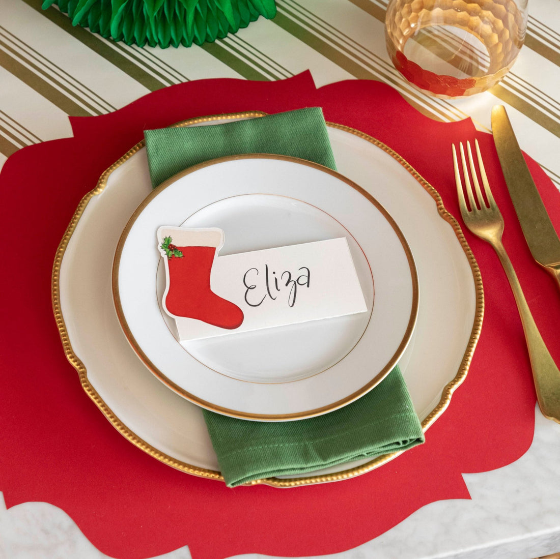 An elegant table setting with the Die-cut Red French Frame Placemat on the Gold Peppermint Stripe Runner, with gold rimmed plates and a a Stocking Place Card with the name &quot;Eliza&quot; written on it, on top.