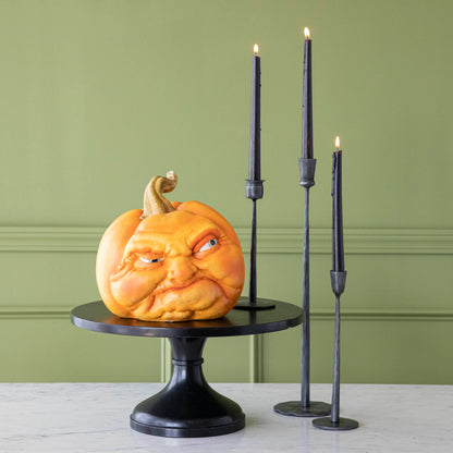 A pumpkin making a mean face, on top of a black pedestal stand surrounded by three black candles.