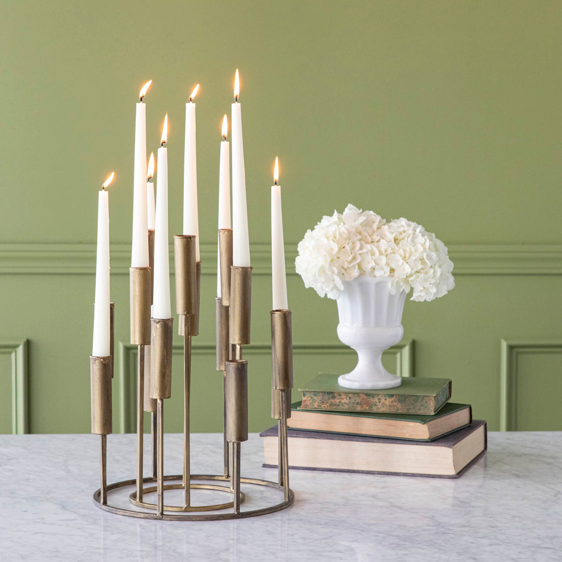 One short iron taper candle holder nestled inside of a tall iron taper candleholder, with various heights of lit candles, on a table with flowers stacked on top of books.