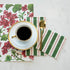 Two Green & Red Awning Stripe Napkins, one Guest and one Cocktail, tucked under a saucer with a cup of coffee on a red and green placemat on a white table, from above.