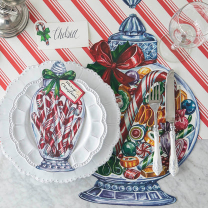 A festive Christmas place setting featuring the Candy Cane Jar Table Accent with &quot;Merry Christmas&quot; written on the tag, resting on dinner plates on top of the Die-cut Candy Jar Placemat.