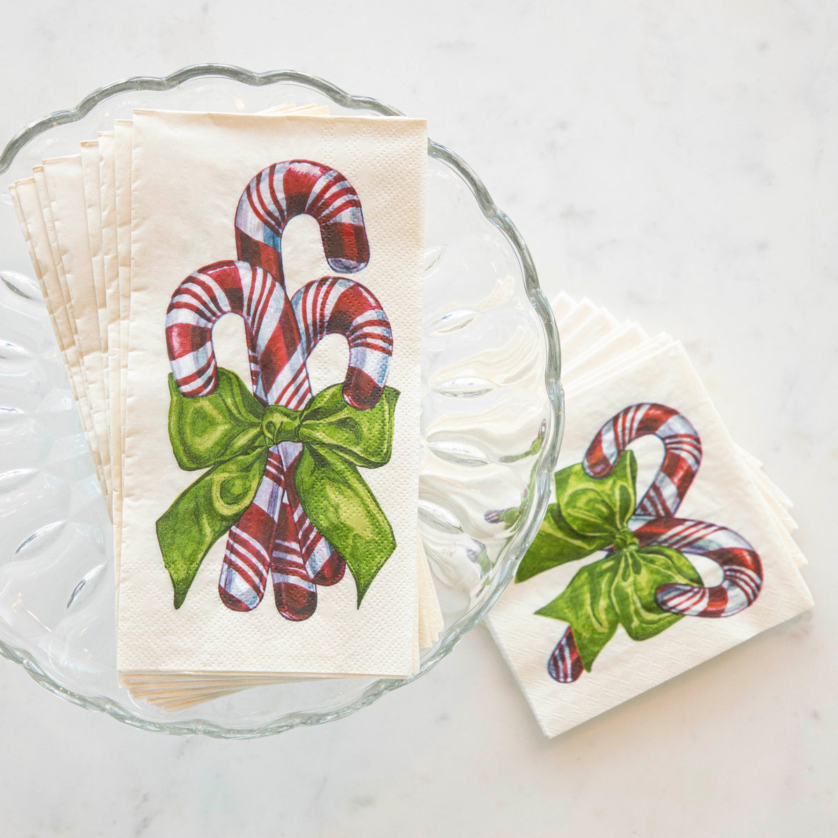 A stack of Candy Cane Guest Napkins fanned out on a glass cake stand, next to a stack of Candy Cane Cocktail Napkins fanned out on the table.