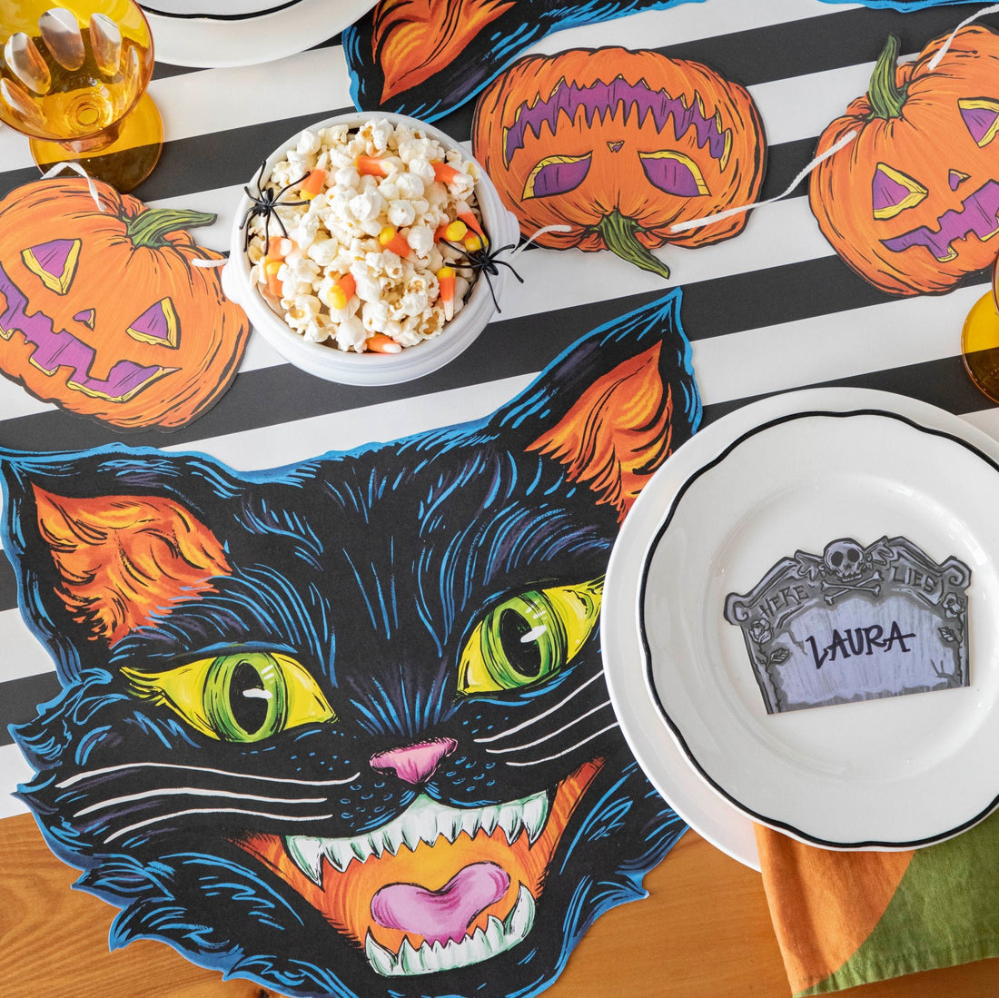  A spooky soiree table setting featuring the Black Classic Stripe Runner, adorned with pumpkin bunting, a bowl of popcorn and candy corn, and a white plate with the Tombstone Place Card labeled &quot;Laura&quot;.