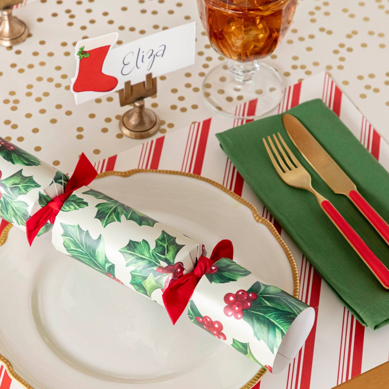 An elegant Christmas themed table setting featuring the Peppermint Stripe Placemat under a gold rimmed dinner plate, green napkin, gold and red silverware, and a Holly Cracker on top.