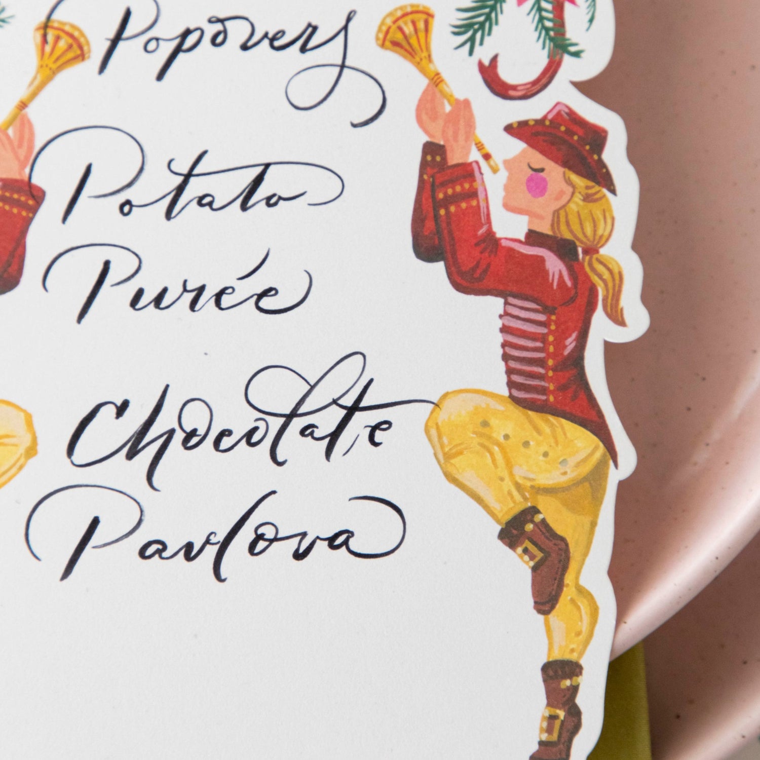 Close-up of a Pipers Piping Table card with a menu written on it resting on the plate of a holiday place setting, showing the illustration of one of the pipers in detail.
