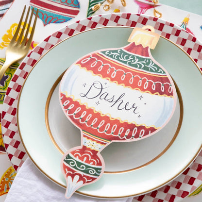 A red Ornaments Table Accent labeled &quot;Dasher&quot; resting on the plate of a Christmas-themed place setting.