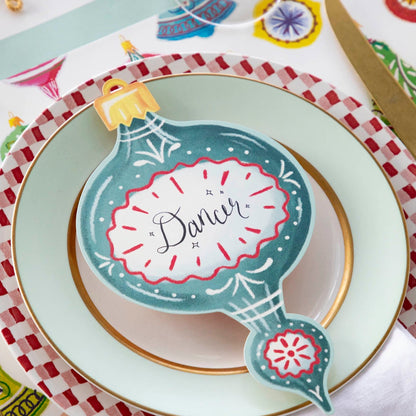 A blue Ornaments Table Accent labeled &quot;Dancer&quot; resting on the plate of a Christmas-themed place setting.