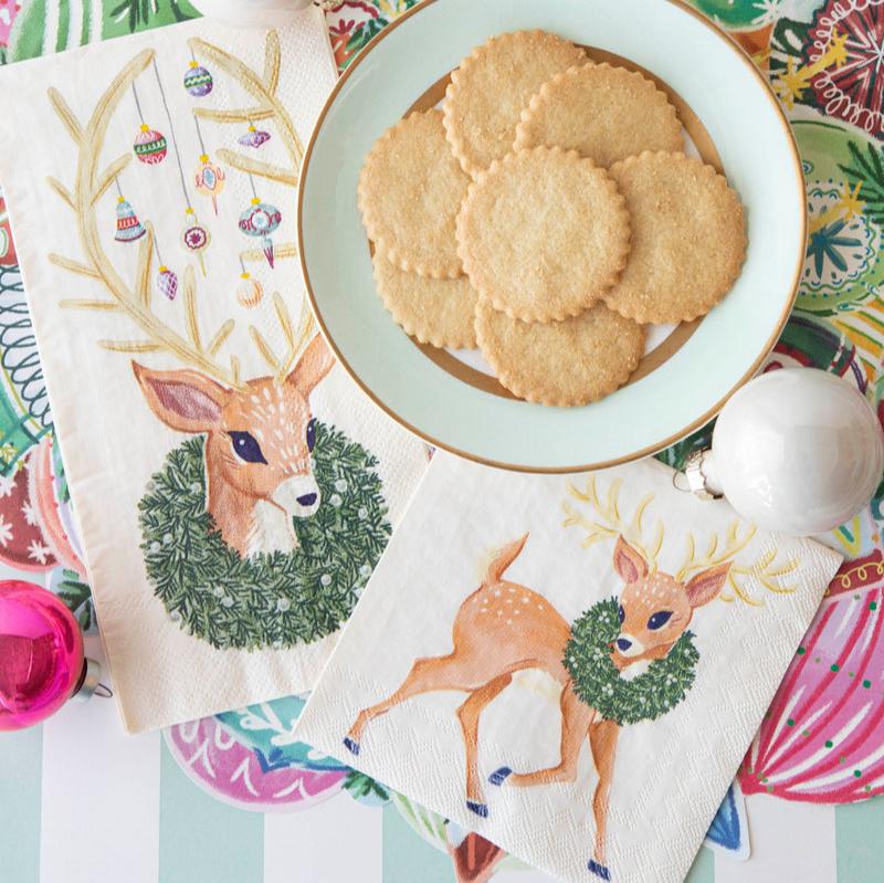 A festive Christmas place setting featuring two Reindeer Napkins, one guest and one cocktail, next to a plate of cookies.