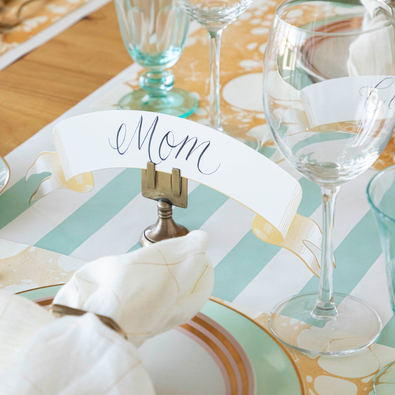 Classic Gold Banner Table Accent with &quot;Mom&quot; written on it, held up by the Brass Place Card holder behind a place setting.